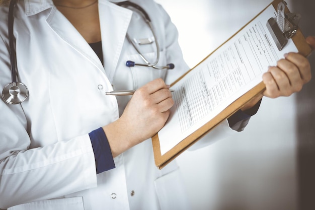 Unknown woman-doctor is writing some notes using a clipboard, while standing in the cabinet in a clinic. Female physician with a stethoscope, close-up. Perfect medical service in a hospital. Medicine