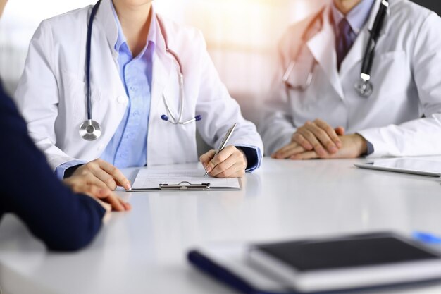 Unknown woman-doctor with male colleague are consulting patient woman while sitting at the desk in sunny clinic, close-up. Covid 2019