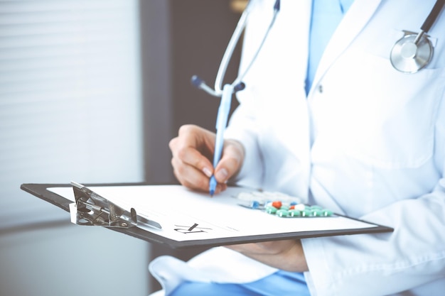 Unknown woman doctor at work at hospital. Pills at medical clipboard. Young female physician write prescription or filling up medical form while sitting in hospital office, close-up.