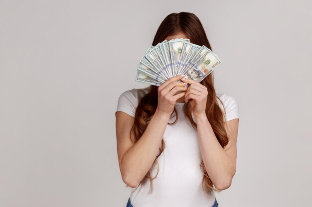 Unknown woman hiding face behind bunch of dollar banknotes, anonymous person holding money, lottery win, big profit, wearing white T-shirt. Indoor studio shot isolated on gray background.