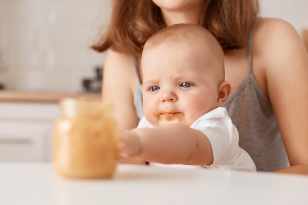 Unknown woman holding feeding infant baby with puree, curious infant stretching hand to jar, family posing in light room at home, childhood and parenthood.