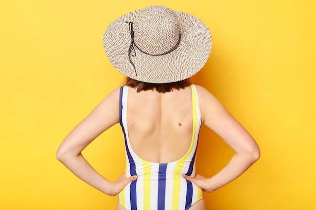 Unknown woman on resort dressed in striped swimwear and straw hat posing isolated over yellow background keeps hands on hips standing backwards to camera showing her back