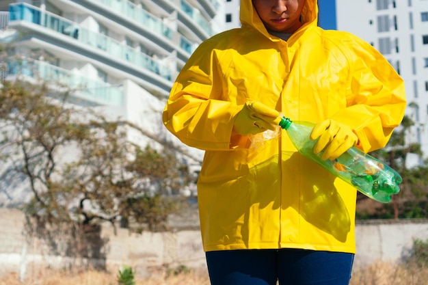 An unknown woman wearing a raincoat and latex gloves, holding a used plastic bottle in her hand, takes care of the planet, cleaning up the park.