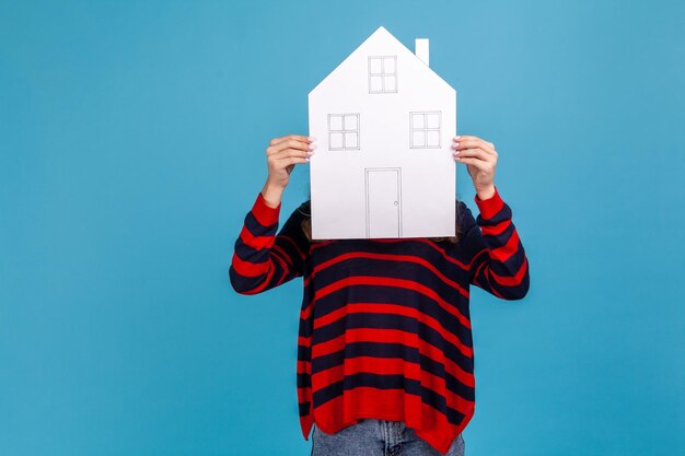 Unknown woman wearing striped casual style sweater, hiding her face behind paper house with drawn windows and door, government housing program. Indoor studio shot isolated on blue background.