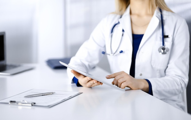 Unknown young woman-doctor is checking some medication names, while sitting at the desk in her cabinet in a clinic. Physician with a stethoscope is using a tablet computer, close-up. Perfect medical s