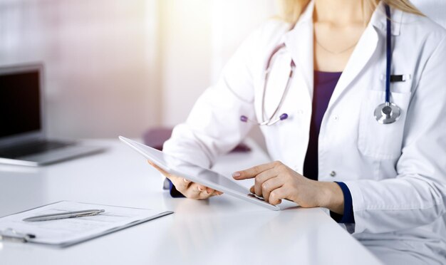 Unknown young woman-doctor is checking some medication names,\
while sitting at the desk in her sunny cabinet in a clinic.\
physician with a stethoscope is using a tablet computer, close-up.\
perfect med