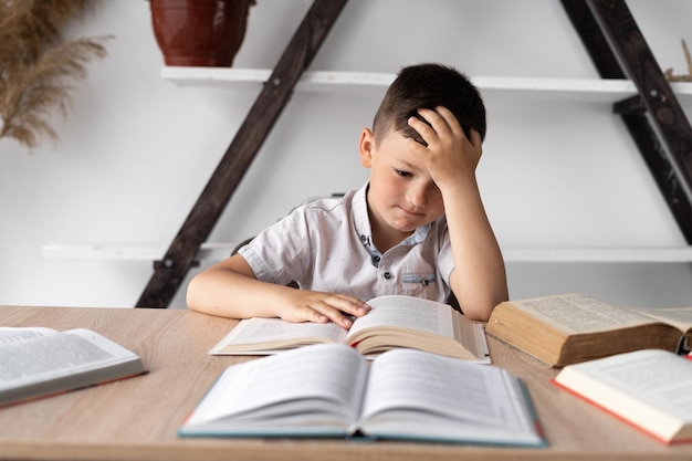 Unmotivated student with headache sitting at home at his desk doing homework preparing for the lesson Tired little school student reading books learning online distance knowledge concept