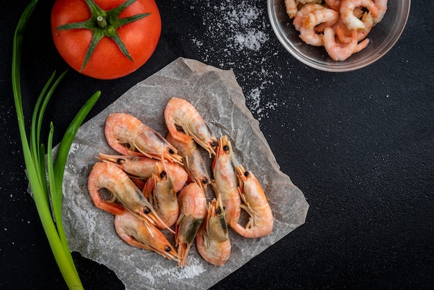 Unpeeled and peeled shrimps with salt, green onion and tomato on black background. Top view.