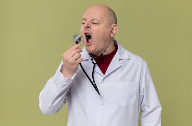 Unpleased adult man in doctor uniform holding and looking at stethoscope 