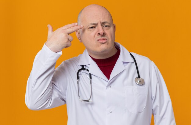 Unpleased adult slavic man in doctor uniform with stethoscope keeping hand at his temple gesturing gun sign