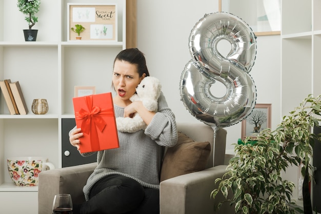 Unpleased beautiful woman on happy women day holding present with teddy bear sitting on armchair in living room