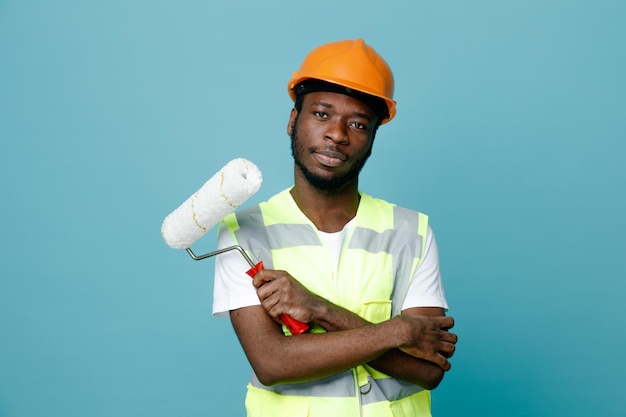 Unpleased crossing hands young african american builder in uniform holding roller brush isolated on blue background