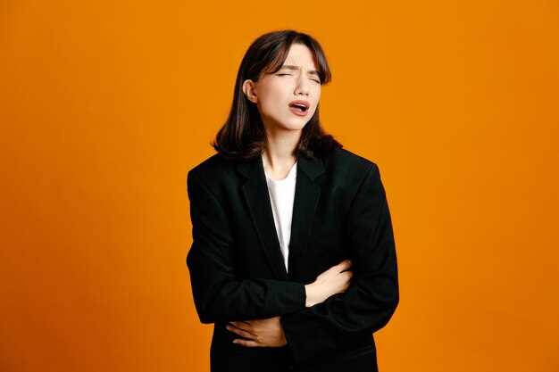 Unpleased grabbed stomach young beautiful female wearing black jacket isolated on orange background