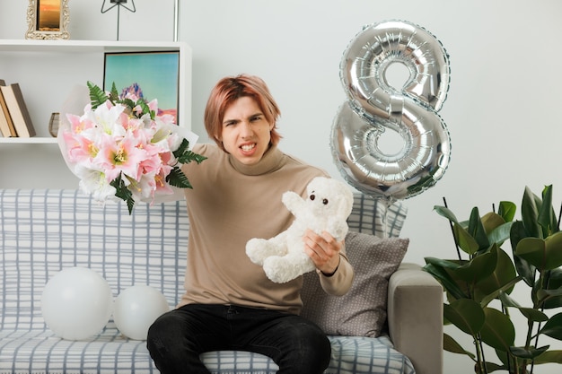 Unpleased handsome guy on happy women day holding bouquet with teddy bear sitting on sofa in living room