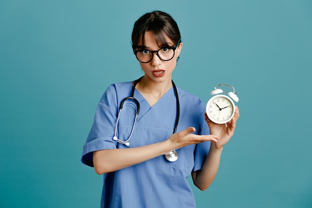 unpleased holding alarm clock young female doctor wearing uniform fith stethoscope isolated on blue background