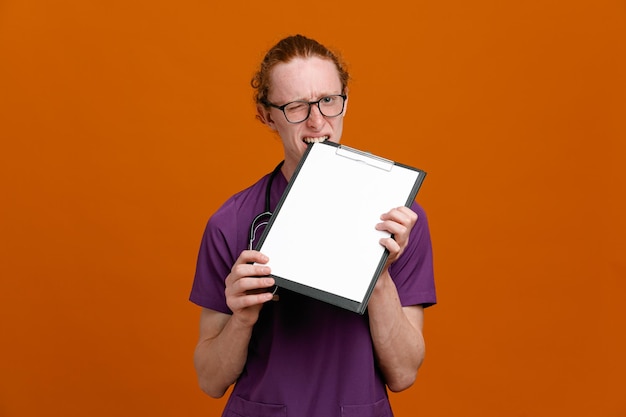 Unpleased holding clipboard in mouth young male doctor wearing uniform with stethoscope isolated on orange background