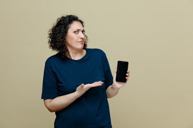 Unpleased middleaged woman wearing tshirt looking at camera showing mobile phone and empty hand isolated on olive green background