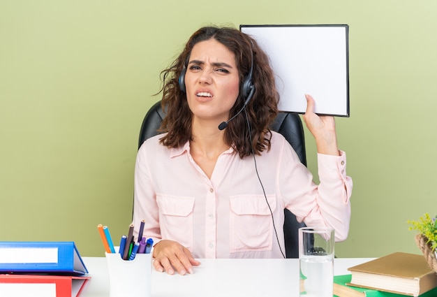 Unpleased pretty caucasian female call center operator on headphones sitting at desk with office tools holding clipboard 