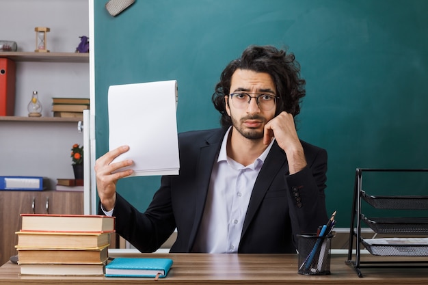 Photo unpleased putting hand on cheek male teacher wearing glasses holding paper sitting at table with school tools in classroom