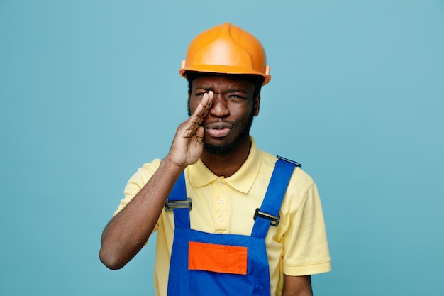 Mettendo dispiaciuto la mano sul viso giovane costruttore afroamericano in uniforme isolato su sfondo blu