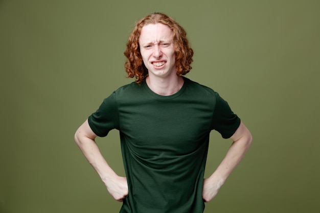 緑の背景に分離された緑のTシャツを着て腰に手を置く不満若いハンサムな男