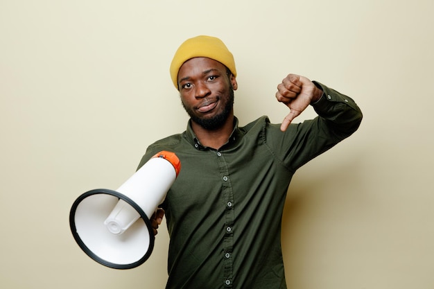 Unpleased showing thumbs down young african american male in hat wearing green shirt holding loudspeaker isoloated on white background