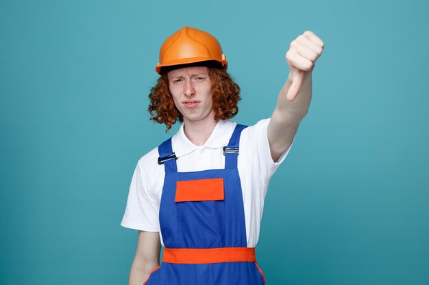 Unpleased showing thumbs down young builder man in uniform isolated on blue background