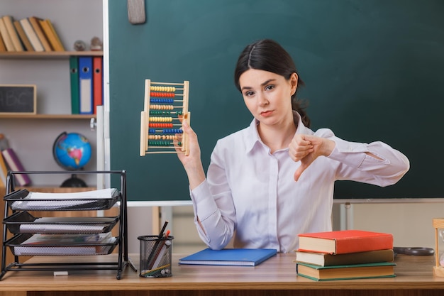 教室で学校の道具を持って机に座っているそろばんを持っている若い女性教師に親指を見せて不満