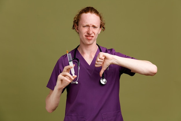 Unpleased showing thumbs down young male doctor wearing uniform with stethoscope holding syringe isolated on green background