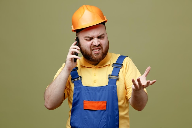 Unpleased spreading hand speaks on the phone young builder man in uniform isolated on green background