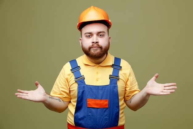 Unpleased spreading hands young builder man in uniform isolated on green background