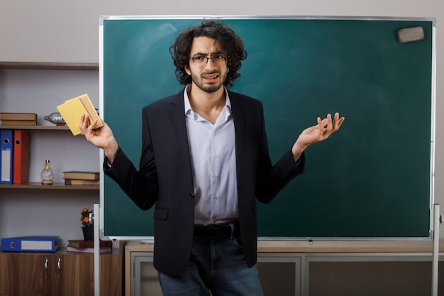 Unpleased spreading hands young male teacher wearing glasses standing in front blackboard holding book in classroom