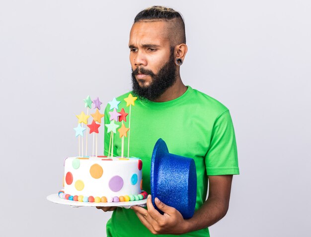 Unpleased young afro-american guy wearing party hat holding cake with party hat 