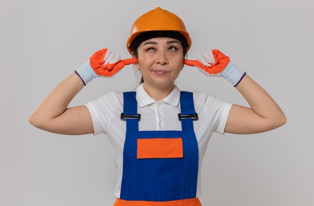 Недовольная молодая азиатская девушка-строитель с оранжевым защитным шлемом и перчатками, закрывающая уши пальцами