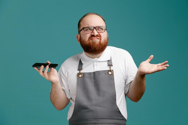 Giovane barbiere scontento che indossa l'uniforme e gli occhiali guardando il lato tenendo il telefono cellulare che mostra la mano vuota isolata su sfondo blu