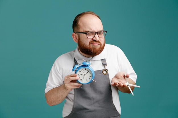 Unpleased young barber wearing uniform and glasses showing holding alarm clock and scissors looking at camera with one eye closed isolated on blue background