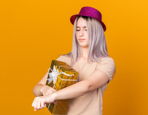 Unpleased young beautiful girl wearing party hat holding gift box showing wrist clock gesture 