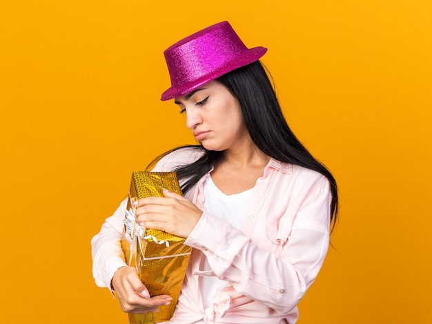 Una giovane bella ragazza scontenta che indossa un cappello da festa che tiene in mano e guarda la confezione regalo