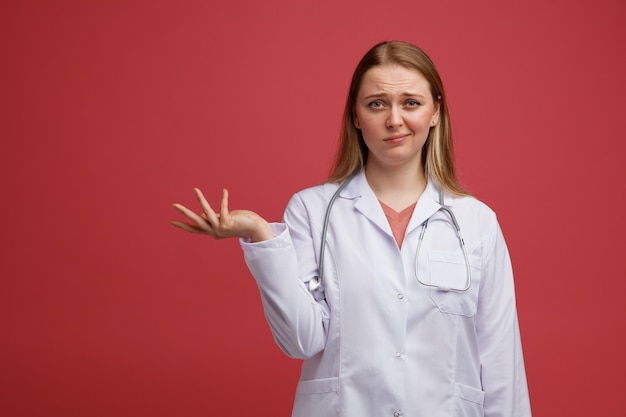 Giovane medico femminile biondo dispiaciuto che indossa veste medica e stetoscopio intorno al collo che mostra la mano vuota