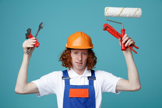 Unpleased young builder man in uniform holding construction tools isolated on blue background