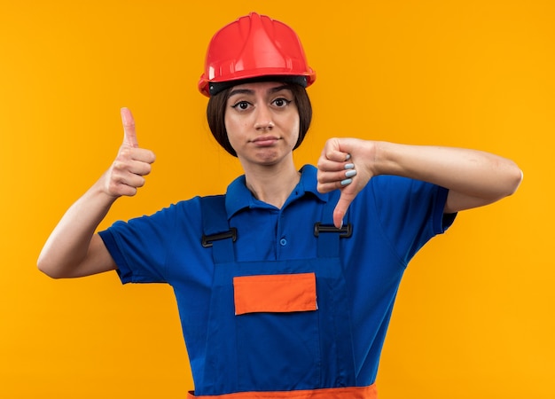 Giovane donna del costruttore scontenta in uniforme che mostra i pollici in su e in giù