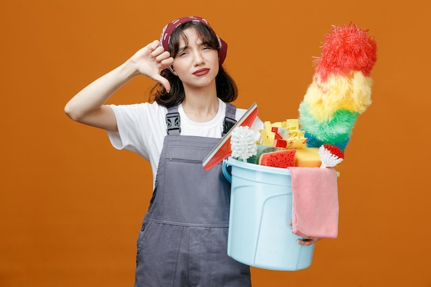 Unpleased young female cleaner wearing uniform and bandana holding bucket full of cleaning tools looking at camera showing thumb down isolated on orange background