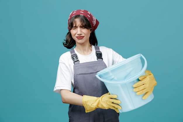 Unpleased young female cleaner wearing uniform bandana and rubber gloves holding bucket with both hands with closed eyes isolated on blue background