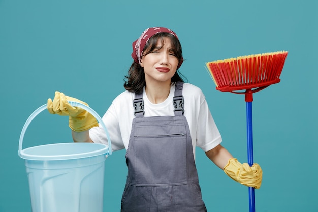 unpleased young female cleaner wearing uniform bandana and rubber gloves holding squeegee mop and bucket looking at camera isolated on blue background