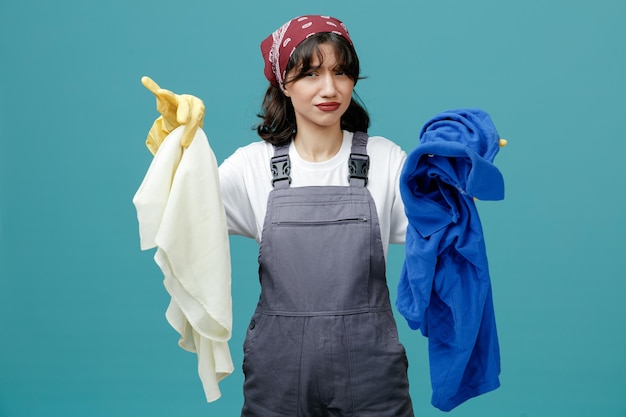 Giovane donna scontenta addetta alle pulizie che indossa bandana uniforme e guanti di gomma guardando la fotocamera che mostra vestiti sporchi isolati su sfondo blu