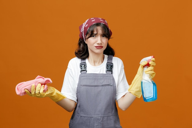 Unpleased young female cleaner wearing uniform rubber gloves and bandana holding cloth duster and cleanser looking at camera isolated on orange background