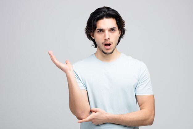 Unpleased young handsome man looking at camera touching elbow showing empty hand isolated on white background