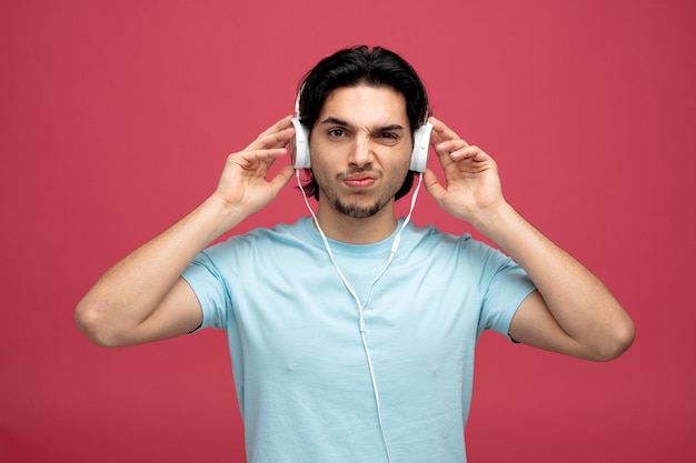 unpleased young handsome man wearing and touching headphones looking at camera isolated on red background