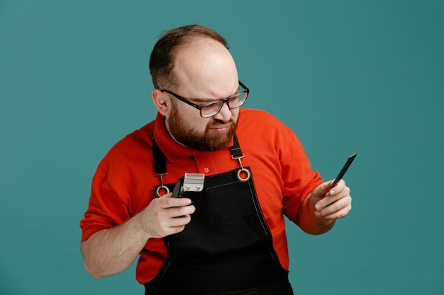 Unpleased young male barber wearing glasses red shirt and barber apron looking down holding hair clipper and teaser comb in air isolated on blue background