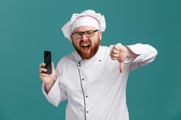 Giovane chef maschio scontento che indossa occhiali uniformi e berretto guardando la fotocamera che mostra il telefono cellulare e il pollice verso il basso isolati su sfondo blu